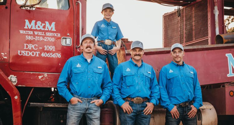 Three adult workers and a young boy from the M&M Water Well Service team pose in front of their red drilling truck. All dressed in vibrant blue uniforms and caps, they display a mix of pride and camaraderie. The boy, standing on the truck, adds a touch of familial warmth to the group portrait at dusk