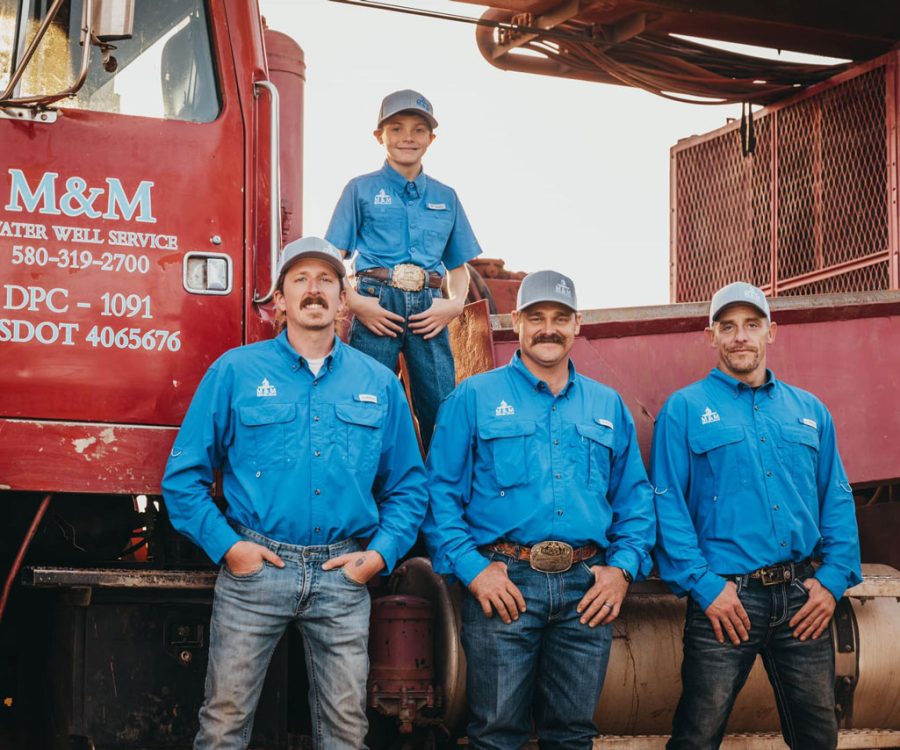 Three adult workers and a young boy from the M&M Water Well Service team pose in front of their red drilling truck. All dressed in vibrant blue uniforms and caps, they display a mix of pride and camaraderie. The boy, standing on the truck, adds a touch of familial warmth to the group portrait at dusk