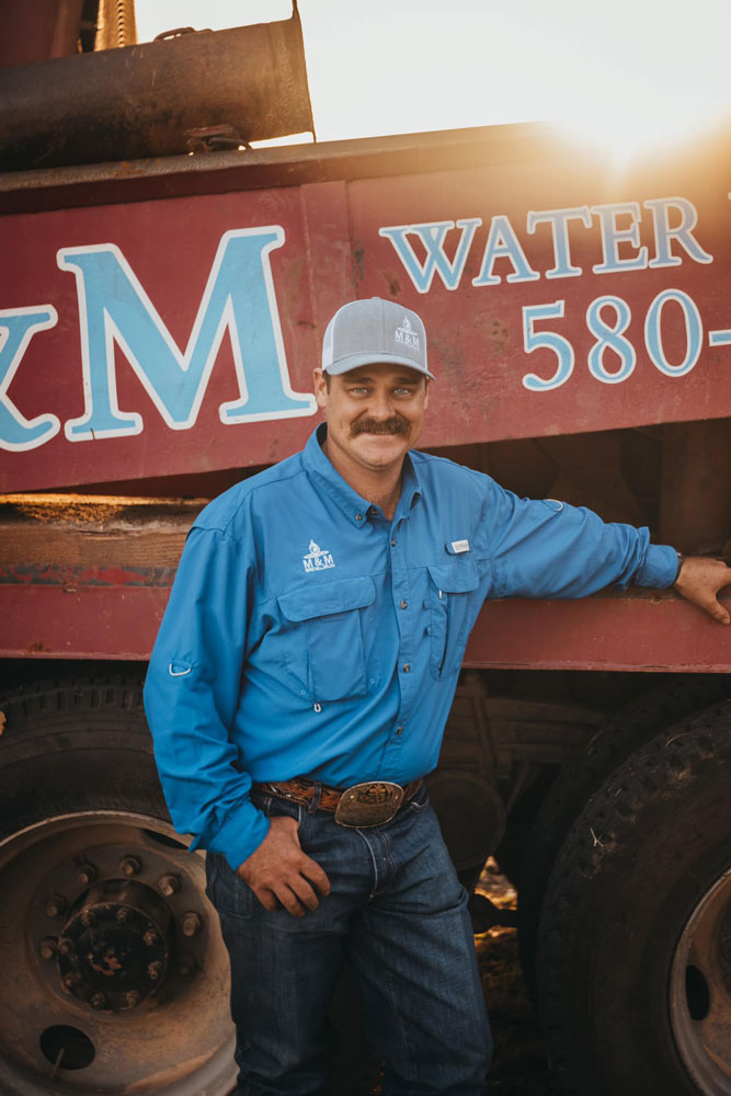 A smiling worker from M&M Water Well Service stands in front of the company's drilling truck, leaning casually against it. He wears a blue uniform shirt, jeans, and a cap, with a backdrop of the truck's logo and contact number. The setting sun casts a warm glow, highlighting his sense of pride and professionalism.