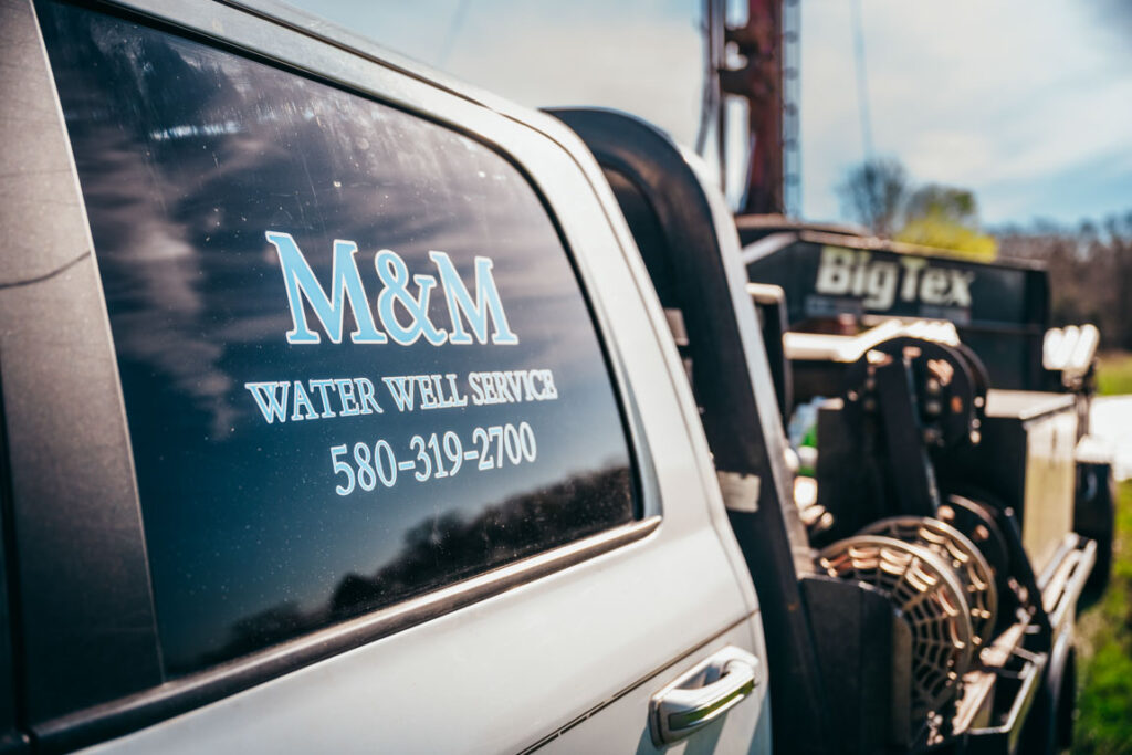 Close-up view of a vehicle door displaying the M&M Water Well Service logo and contact number, '580-319-2700,' with a reflection of the sky visible on the window. The image highlights the professional branding and accessibility of the water well service company, with equipment visible in the background.
