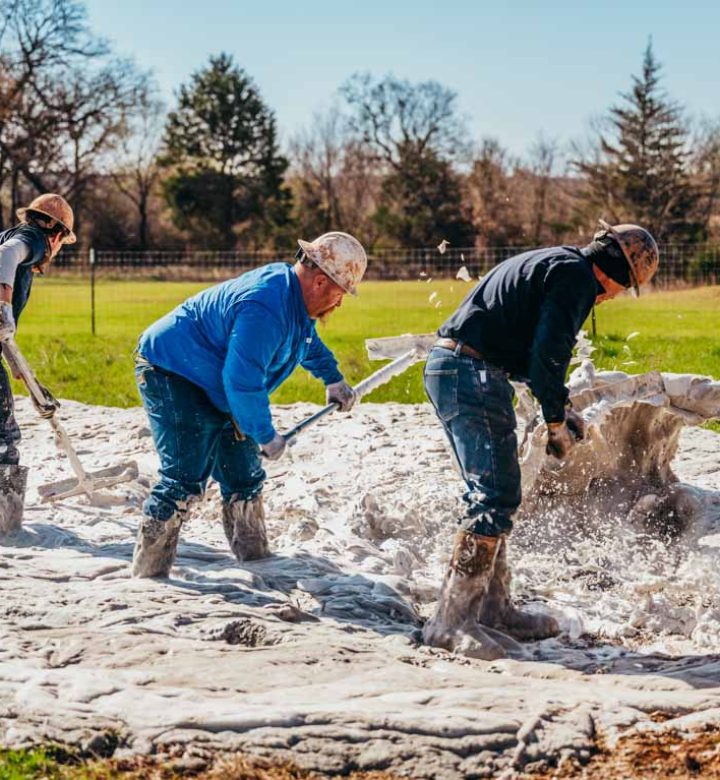 Three workers in protective gear, including hard hats and boots, manage drilling mud at a water well site. They use shovels and other tools to handle the thick, wet material spread across the ground. The site is surrounded by grass and trees, with a clear sky overhead.