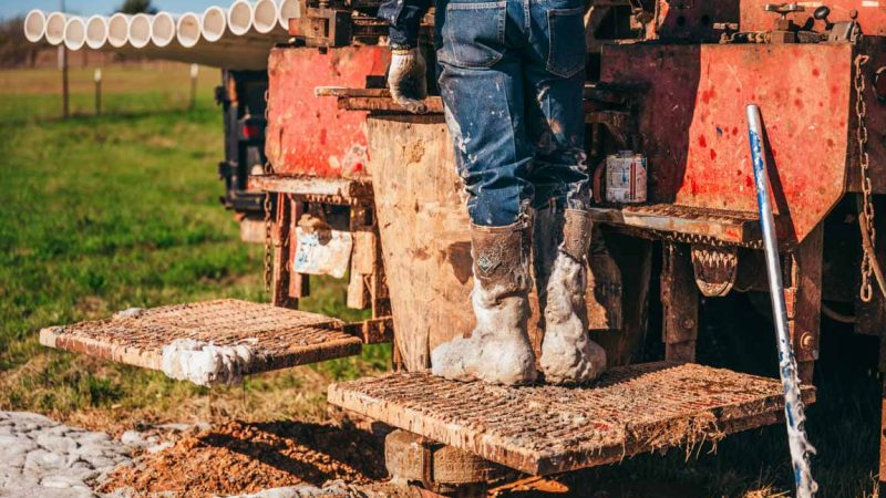 A close-up of a worker's muddy boots standing on the metal steps of a well-used drilling rig. In the background, rows of PVC pipes are lined up, ready for insertion into the drilled well. The image captures the rugged conditions and essential equipment of a water well drilling operation.
