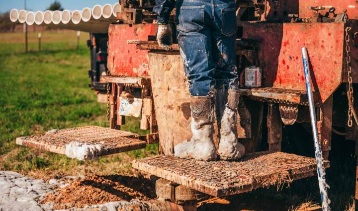 A close-up of a worker's muddy boots standing on the metal steps of a well-used drilling rig. In the background, rows of PVC pipes are lined up, ready for insertion into the drilled well. The image captures the rugged conditions and essential equipment of a water well drilling operation.