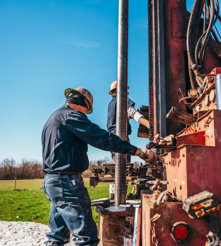 Two workers operate a large drilling rig, managing the steel drill pipe extending into the ground. Dressed in work gear and engaged in manipulating complex machinery, they exemplify the skilled labor involved in drilling for water in a rural environment under a clear blue sky.
