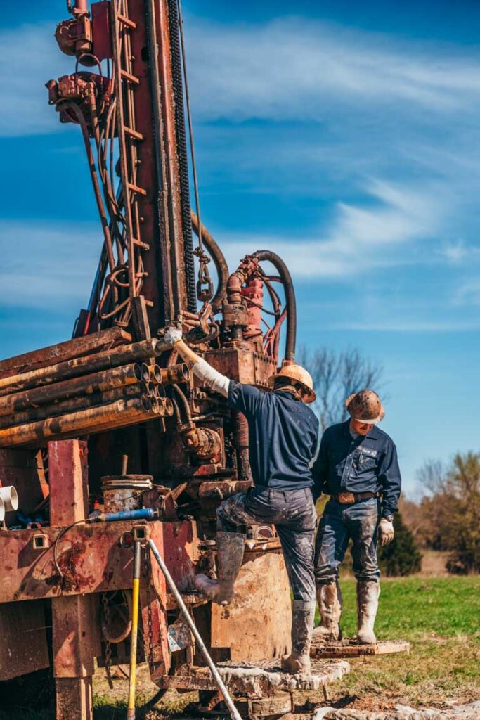 Two workers engage in precise adjustments on a large drilling rig. One stands on the platform, manipulating the rig's controls, while the other observes. The machinery, marked by heavy use, highlights the technical and labor-intensive nature of water well drilling, set against a partly cloudy rural backdrop.