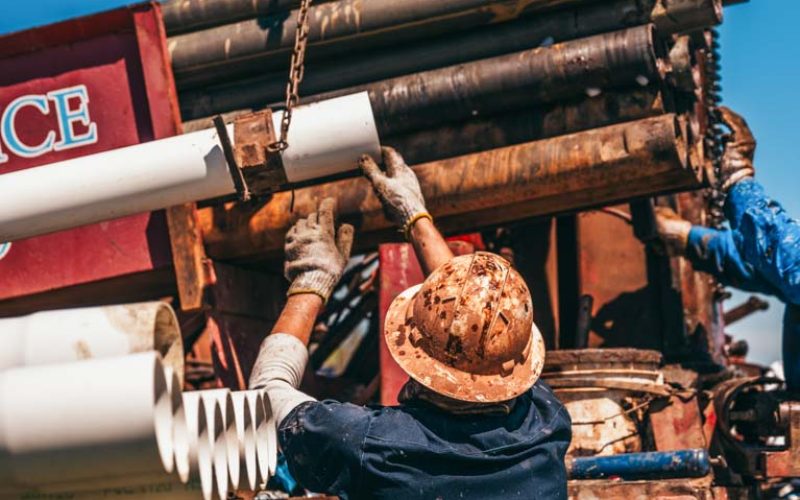 A worker in a hard hat and gloves lifts a PVC pipe towards the drilling rig, ready to install it. The scene captures the precision and effort required in the well drilling process, with various pipes and machinery in the background. The bright, clear day highlights the demanding nature of the task.