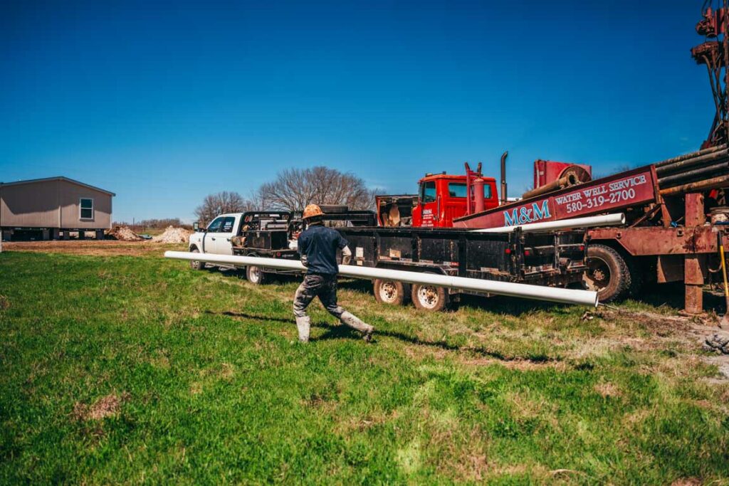A worker carries a long PVC pipe across a grassy field towards a well drilling rig, marked with the company name 'M&M Water Well Service.' The rural backdrop features a mobile home and several vehicles, illustrating the logistics and manual effort involved in setting up a water well.