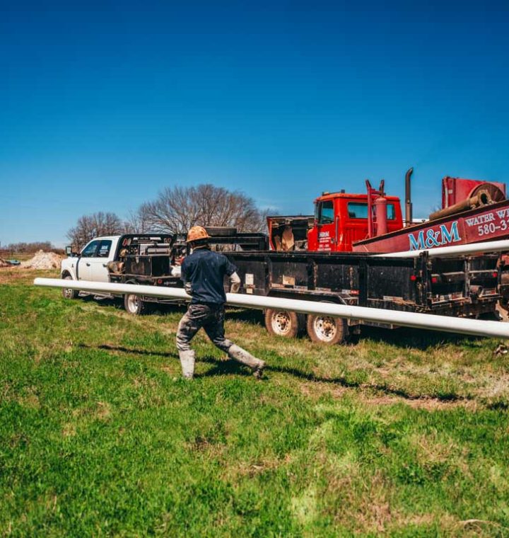 A worker carries a long PVC pipe across a grassy field towards a well drilling rig, marked with the company name 'M&M Water Well Service.' The rural backdrop features a mobile home and several vehicles, illustrating the logistics and manual effort involved in setting up a water well.