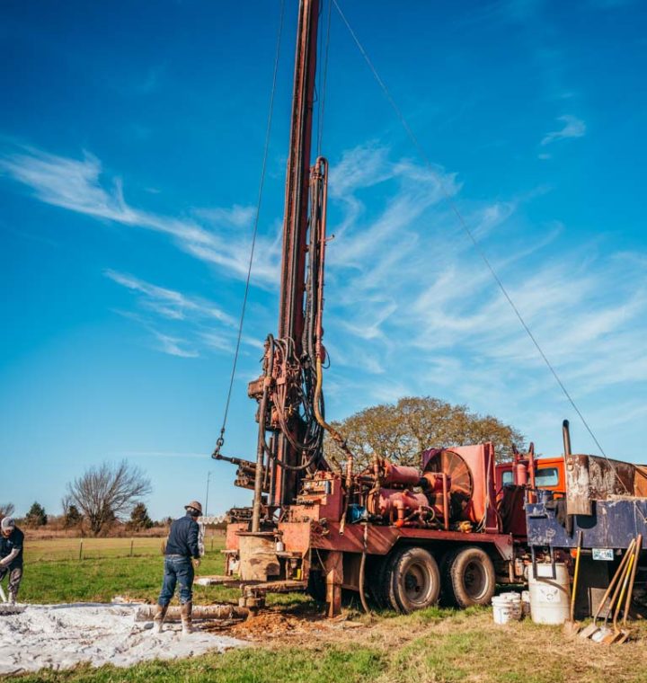 A tall water well drilling rig operates in an open field, with a worker overseeing the process near the base of the rig. The scene shows various tools and equipment spread around, set against a clear blue sky with scattered clouds. The backdrop includes green grass and distant trees, illustrating a typical day of drilling operations.