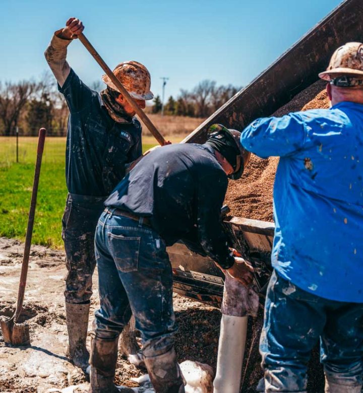 Three workers at a well drilling site handle equipment and materials with coordinated effort. One uses a shovel to manage the overflow of materials, another adjusts machinery, and the third oversees the operation. Their mud-covered workwear and focused expressions highlight the intensive labor involved in installing a water well under a clear sky.