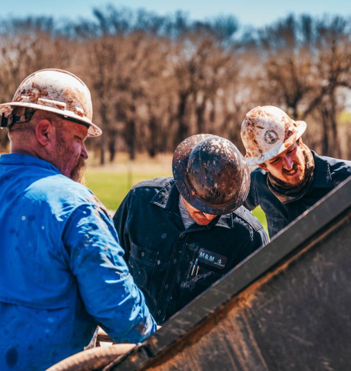 Three well drilling workers, deeply engaged in a collaborative task, examine equipment together. Dressed in rugged work gear and hard hats speckled with mud, they represent the teamwork essential in navigating the complexities of rural well drilling. The lush tree line in the background underscores the remote and natural work setting.