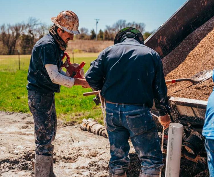 Two workers at a well drilling site, one holding a measurement tool, focusing intently on their task. They are covered in mud, illustrating the messy conditions of outdoor manual labor. The rural landscape in the background and equipment scattered around emphasize the challenging environment of their work.