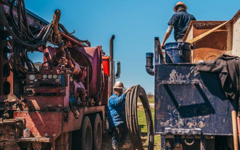 Two workers actively engaged in a water well drilling operation in a rural area, surrounded by specialized machinery. One worker stands atop a red truck adjusting equipment, while the other manages heavy hoses on the ground, indicative of the process to access underground water sources.