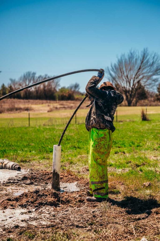 A worker, clad in bright yellow pants and a mud-covered black jacket, maneuvers a large hose at a water well drilling site. He stands with the hose elevated, surrounded by green fields and trees under a clear blue sky, showcasing the hands-on effort involved in water well drilling operations.