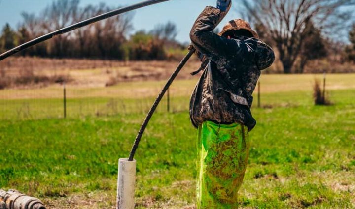 A worker, clad in bright yellow pants and a mud-covered black jacket, maneuvers a large hose at a water well drilling site. He stands with the hose elevated, surrounded by green fields and trees under a clear blue sky, showcasing the hands-on effort involved in water well drilling operations.
