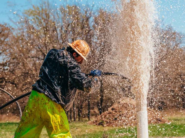 A worker in vivid yellow pants and a mud-splattered black jacket manages a high-pressure eruption of water from a newly drilled well. He uses a hose to direct the gushing water, creating a spectacular splash against the backdrop of trees and a clear sky, illustrating the dynamic nature of water well drilling.