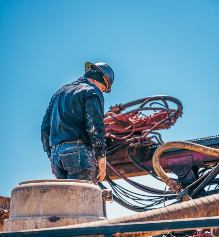 A water well driller, seen from behind, works attentively on top of a large drilling machine. The worker is dressed in a dark jacket and jeans, with a safety helmet on. Intricate machinery with hoses and metal components forms the backdrop under a clear blue sky.