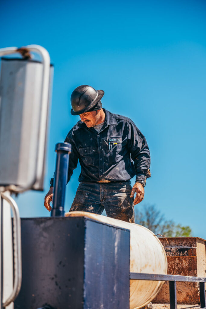 A focused water well driller, dressed in a black uniform and hard hat, stands on a drilling truck. He is managing a large cylindrical piece of equipment, possibly preparing for a drilling operation. The clear blue sky in the background enhances the outdoor work environment.