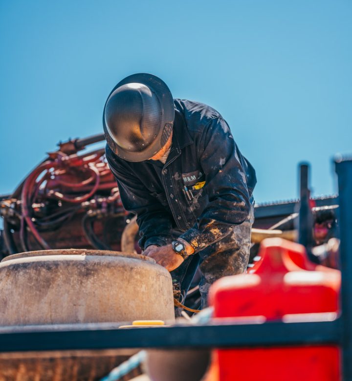 A worker in a black jacket and camouflage pants is focused on repairing a piece of water well drilling equipment. The worker, wearing a hard hat and gloves, is adjusting parts on the machinery, which is mounted on a truck. A clear blue sky serves as the background.