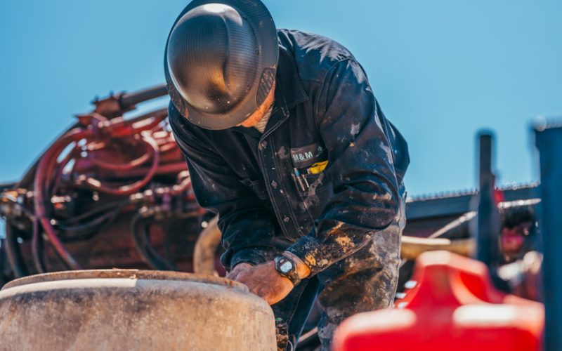 A worker in a black jacket and camouflage pants is focused on repairing a piece of water well drilling equipment. The worker, wearing a hard hat and gloves, is adjusting parts on the machinery, which is mounted on a truck. A clear blue sky serves as the background.