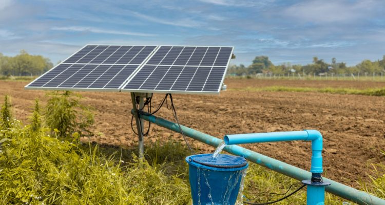 Image of a solar-powered well system in a field, featuring a set of solar panels mounted on a metal frame and a blue pipe delivering water into a blue container. The system is set against a backdrop of tilled soil and greenery, with a clear blue sky above.